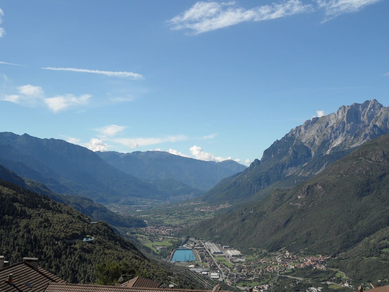 Il fondovalle della Valle Camonica visto dall'alto: capannoni, un bacino e la linea ferroviaria sul piano, i paesi distribuiti sui versanti boscosi.